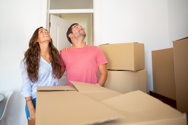 Взволнованная счастливая пара смотрит на свою новую квартиру, стоит среди стопок картонных коробок и обнимается