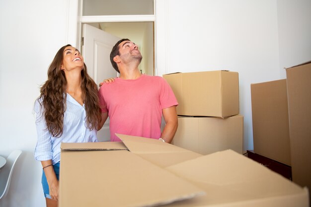 Excited joyful couple looking over their new apartment, standing among stacks of carton boxes and hugging