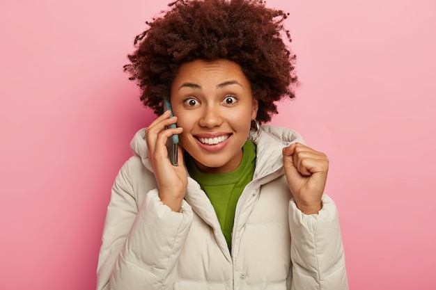 Free photo excited joyful dark skinned woman raises clenched fist, chats on smartphone, discusses details of meeting, makes telephone conversation, dressed in warm winter outerwear, looks gladfully at camera