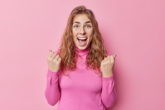 Excited joyful European woman exclaims yes clenches fists triumphs over something celebrates success wears turtleneck isolated over pink background being supportive and happy about something