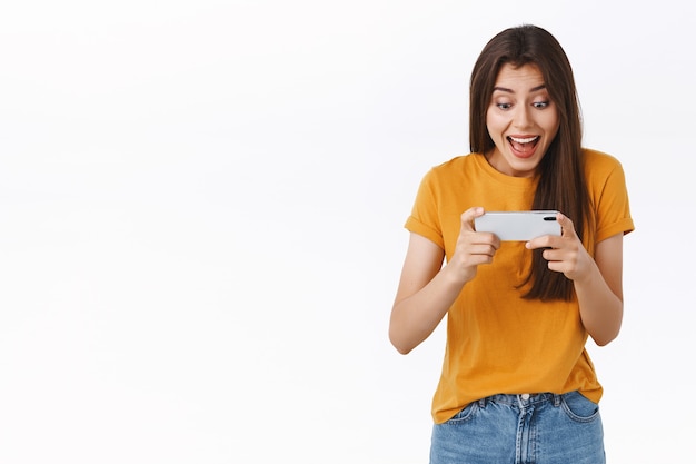 Excited, joyful good-looking woman in yellow t-shirt playing awesome amusing new mobile game, holding smartphone horizontally, cheering, smiling and looking phone display astonished