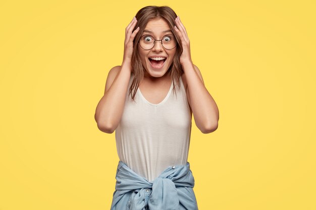 Excited joyful lady has overjoyed expression, gazes with carefree expression, keeps both hands on head, exclaims with happiness, wears casual vest, shirt around waist, poses indoor