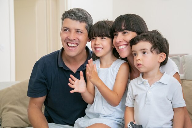 Excited joyful parent couple with two kids watching TV, sitting on couch in living room, looking away and smiling.