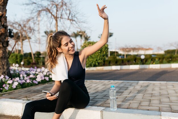 Excited joyful young woman in sportswear gratulating to side in sunny morning. Expressing positivity, sitting on street of tropical city, smiling, training, healthy lifestyle.