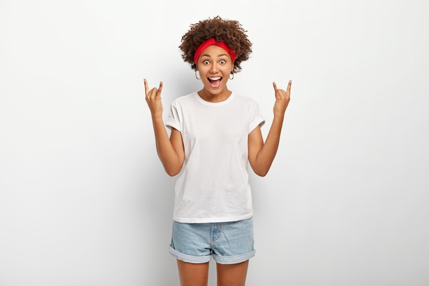 Excited joyous dark skinned female looks with joy at camera, shows rock n roll heavy metal gesture, dressed in casual outft, isolated over white background. Body language.