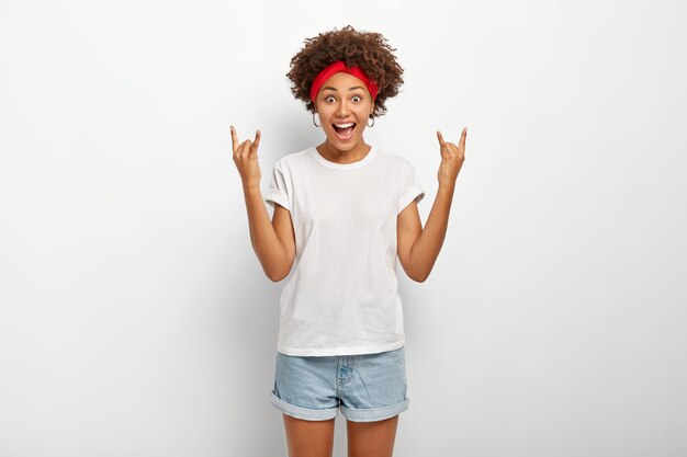 Excited joyous dark skinned female looks with joy at camera, shows rock n roll heavy metal gesture, dressed in casual outft, isolated over white background. Body language.