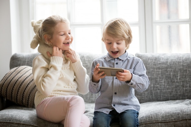 Free photo excited kids having fun using smartphone sitting together on sofa