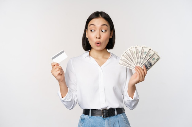 Excited korean girl looking at credit card holding money cash posing against studio background