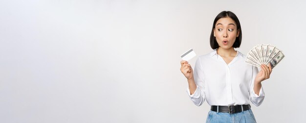 Excited korean girl looking at credit card holding money cash posing against studio background
