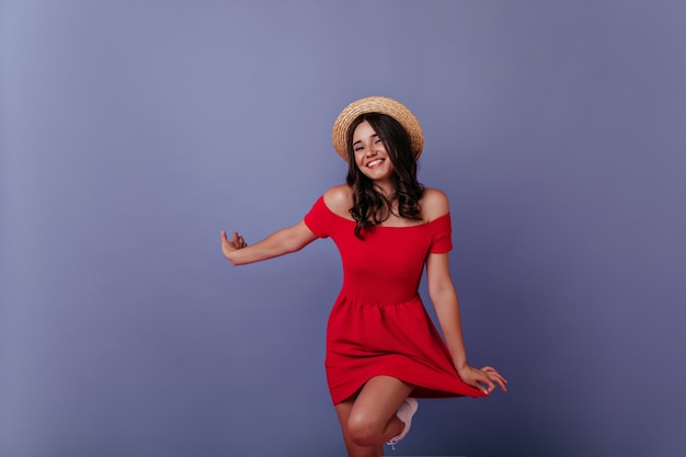 Excited lady in elegant boater posing . Amazing caucasian girl in red dress isolated on purple wall.