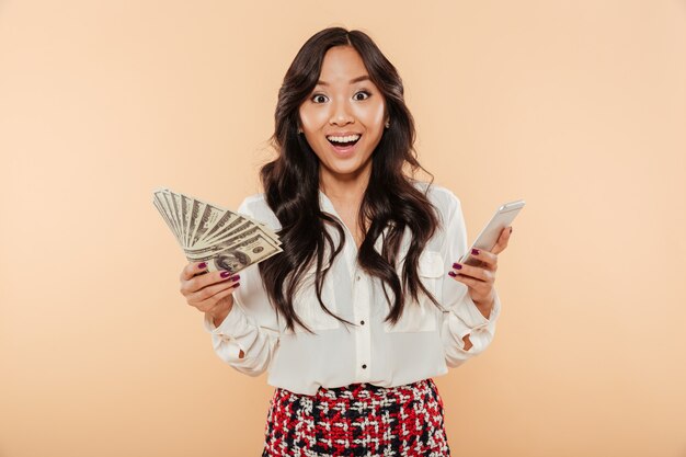 Excited lady holding fan of 100 dollar bills in one hand and trendy smartphone in another being shocked with huge amount of money over peach background