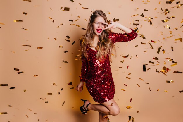 Возбужденная дама с модным макияжем забавно позирует под сверкающим конфетти. Замечательная молодая женщина в стильном красном платье танцует с улыбкой на бежевом фоне на вечеринке.