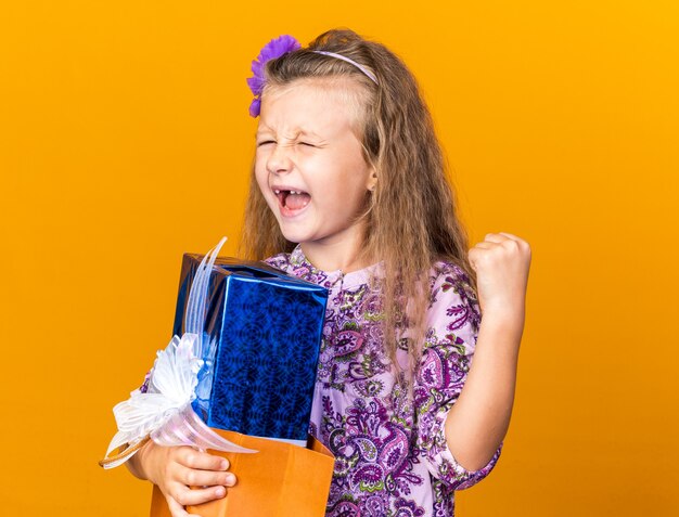 흥분된 작은 금발 소녀 선물 상자를 들고 복사 공간이 오렌지 벽에 고립 된 주먹을 유지
