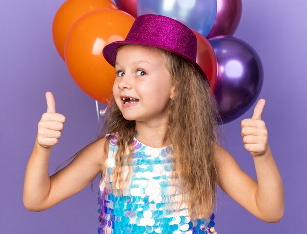 excited little blonde girl with violet party hat thumbing up standing in front of helium balloons isolated on purple wall with copy space