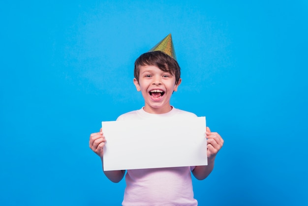 Excited little boy wearing party hat holding blank card in hand on blue surface