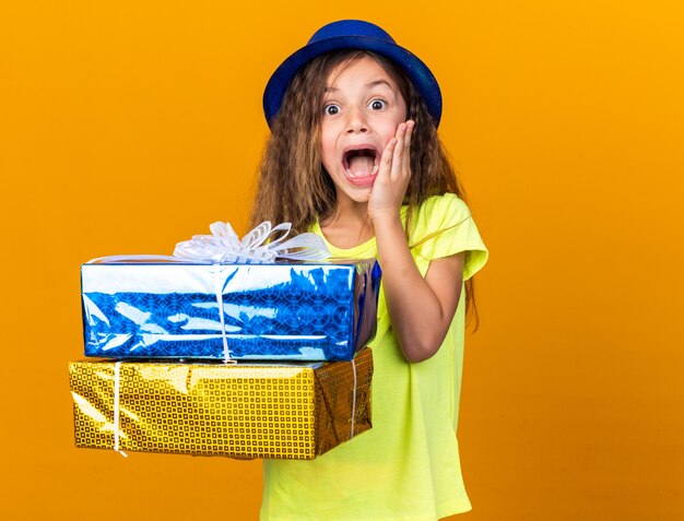 파란색 파티 모자 얼굴에 손을 넣고 복사 공간이 오렌지 벽에 고립 된 선물 상자를 들고 흥분된 어린 백인 소녀