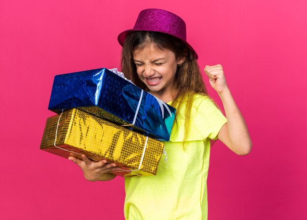 excited little caucasian girl with purple party hat standing with closed eyes keeping fist up and holding gift boxes isolated on pink wall with copy space