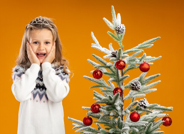 Excited little girl standing nearby christmas tree wearing tiara with garland on neck putting hands on cheeks isolated on orange background