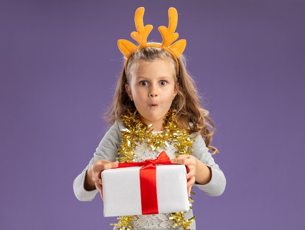Excited little girl wearing christmas hair hoop with garland on neck holding out gift box  isolated on blue wall