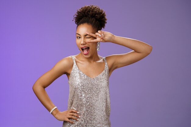 Excited lively good-looking african woman in silver shiny evening dress having fun party celebrating show lucky peace or victory gesture near eye winking happily smiling, standing blue background.