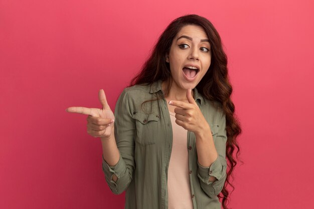 Excited looking at side young beautiful girl wearing olive green t-shirt points at side isolated on pink wall with copy space