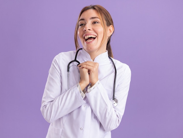 Excited looking at side young female doctor wearing medical robe with stethoscope holding hands together isolated on purple wall