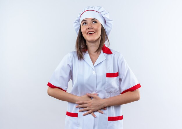 excited looking up young female cook wearing chef uniform grabbed stomach isolated on white wall