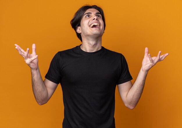 Excited looking up young handsome guy wearing black t-shirt spreading hands isolated on orange wall