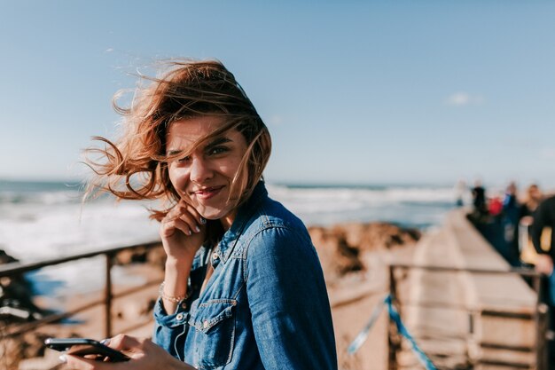 笑顔で屋外の写真撮影を楽しんでいる興奮した愛らしいモデル海の岸で音楽を聴いてポーズをとる幸せな女性