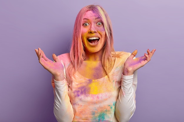 Excited lovely female laughs happily, raises hands, has multicolored face covered with powder paint during Holi festival, celebrates Indian holiday and arrival of spring, models over purple wall