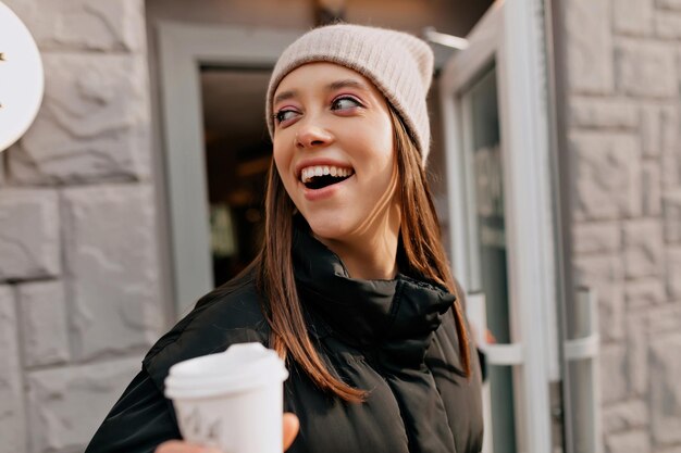 晴れた暖かい春の日に街を歩いているかなり魅力的な女性が行くためにコーヒーと一緒にカフェから出てくるニット帽の大きな笑顔で興奮した素敵な女の子