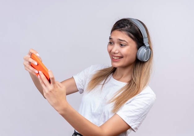 An excited lovely young woman in white t-shirt in headphones playing game on mobile phone