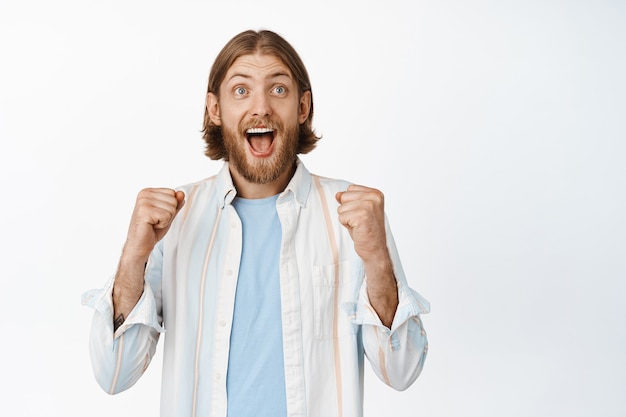 Free photo excited lucky man, winning and celebrating, raising fists up and screaming, rooting for team, become champion, triumphing, standing in shirt on white