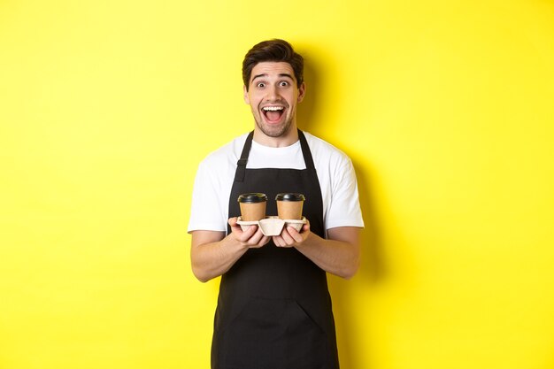 テイクアウトコーヒー2杯を保持し、カフェで働いて、黄色の背景の上に立っている興奮した男性のバリスタ。