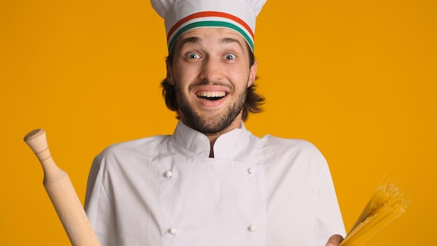 Excited male chef dressed in uniform holding wood rolling pin and pasta looking surprised at camera over yellow background Young man in chef hat looking inspired