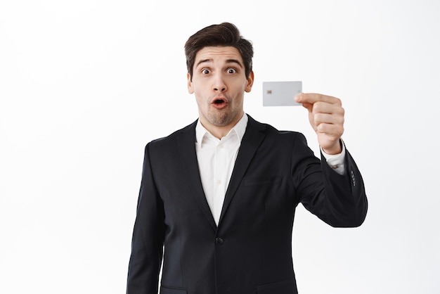 Excited male entrepreneur corporate man shows credit card with wow face impressed by bank offer standing against white background Copy space