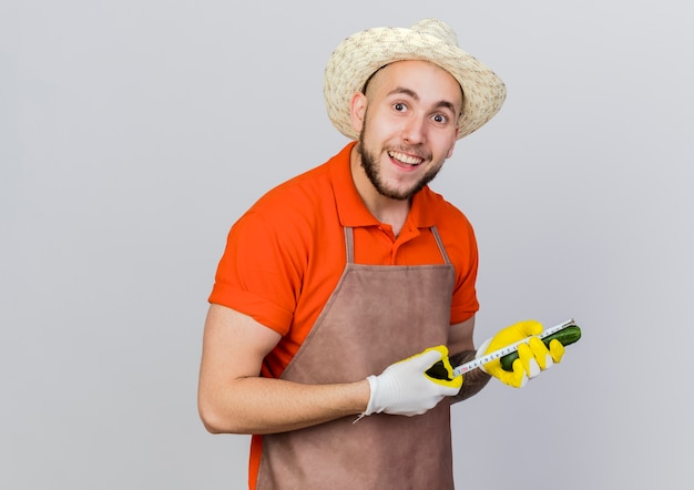 Excited male gardener wearing gardening hat measures cucumber with tape measure 