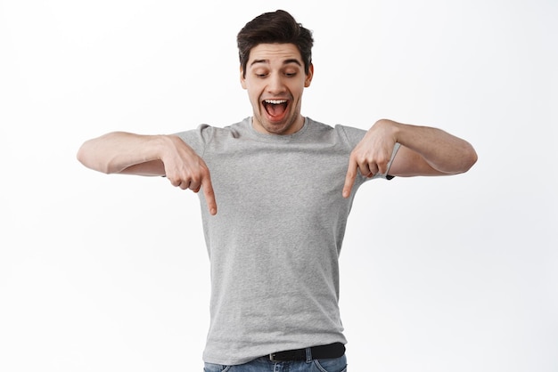 Free photo excited male model in tshirt pointing and looking down shouting yes and smiling amazed found cool thing showing advertisement bellow standing over white background