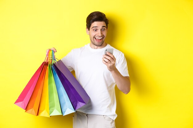 Excited man holding shopping bags and looking happy at mobile phone screen, standing over yellow background.