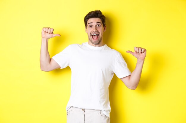 Excited man looking happy, pointing at himself with amazement, standing over yellow background.