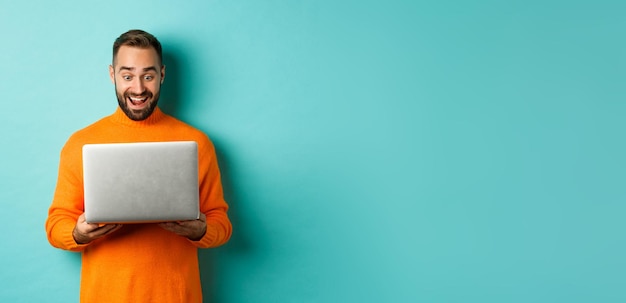 Uomo eccitato in maglione arancione che lavora al computer portatile guardando lo schermo del computer stupito in piedi sopra il ligh