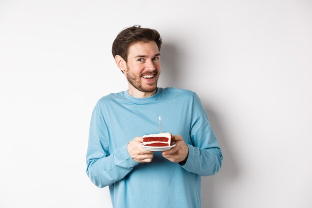 Excited man receive birthday surprise, holding bday cake and smiling happy, standing over white background, making wish on lit candle.