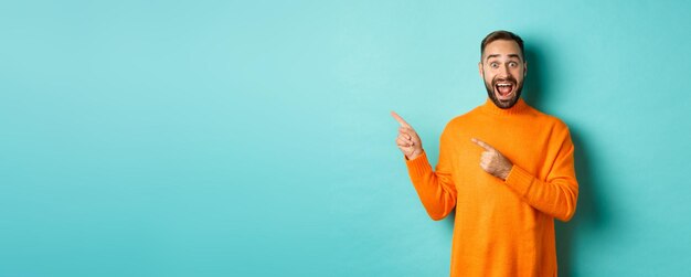 Excited man showing amazing promo offer pointing fingers left at copy space on turquoise background