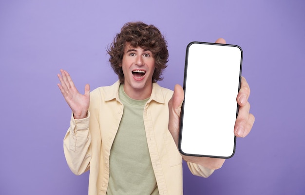 Excited man showing smartphone mockup of blank screen with open mouth and spreading hands isolated on purple background mobile phone mockup for social media application