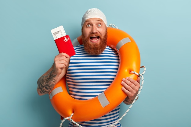 Free photo excited man shows flying boarding pass tickets with passport, carries orange lifebuoy, prepares for trip abroad