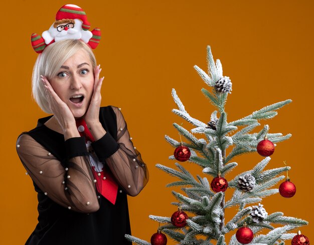 Excited middle-aged blonde woman wearing santa claus headband and tie standing near decorated christmas tree keeping hands on face  isolated on orange wall