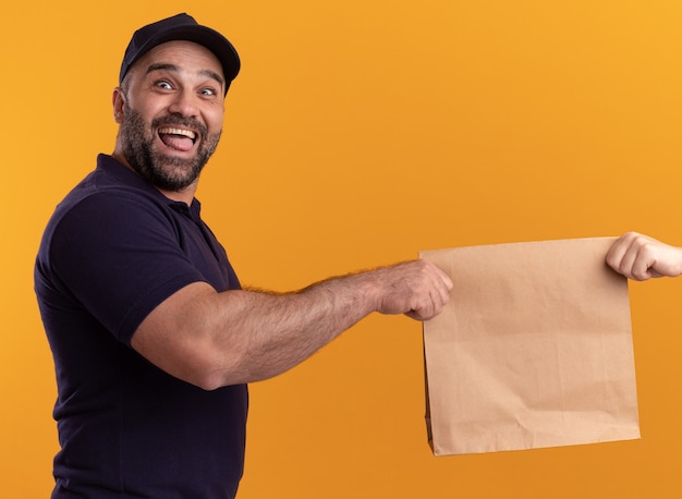 Free photo excited middle-aged delivery man in uniform and cap giving paper food package to client isolated on yellow wall