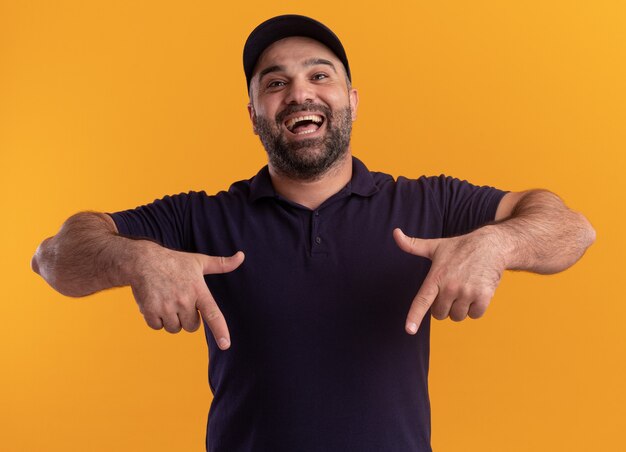 Excited middle-aged delivery man in uniform and cap points at down isolated on yellow wall