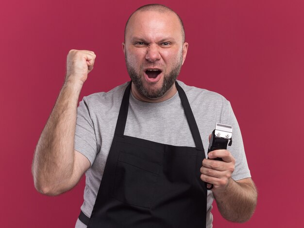 Excited middle-aged male barber in uniform holding hair clippers showing yes gesture isolated on pink wall