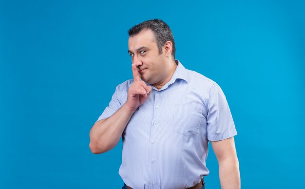 Excited middle-aged man in blue shirt showing shhh sign finger to lips on a blue space